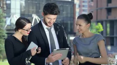 Üç dinamik iş adamı, bir adam ve iki kadın, açık havada neşeli bir anı paylaşıyor, gülümsüyor ve bir tabletle meşgul oluyorlar. Şık kıyafetleri ve canlı ifadeleri modern bir ofise karşı kurulan bir başarı ve takım çalışması hissini yansıtıyor.