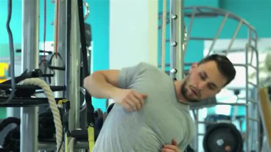 Güçlü bir vücudu olan odaklanmış genç bir adam hareketli bir spor salonunda dinamik bir omuz kemeri esneme rutinine başlar. Kararlı ifadesi ve atletik kıyafeti, aktif bir sporcunun enerjisini sergileyerek spor ve sağlık konusundaki kararlılığını vurguluyor.