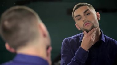 Şık bir berber dükkanında genç bir adam, modern tarz ve klasik tımarın bir karışımını sergileyerek sakalını özenle inceliyor. Atmosfer canlı ve davetkar, parlak aletler ve parlak bir ayna onun yakışıklı özelliklerini yansıtıyor.