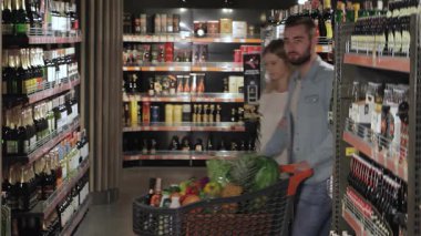 Neşeli bir çift hareketli bir hipermarkette renkli alkollü içecekler arasından zevkle seçim yapıyor. Genç beyaz erkek ve kadın, ışıl ışıl gülümserken, taze ürünlerle dolu alışveriş arabalarına sarılıp,