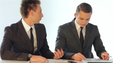 İki genç erkek iş adamı parlak bir masada otururken kağıt üzerinde bir iş planını analiz ederek odaklanmış bir tartışmaya giriştiler. Özenli ifadeleri ve profesyonel kıyafetleri, bir şirkette takım çalışmasının ve stratejik düşüncenin önemini vurgular.