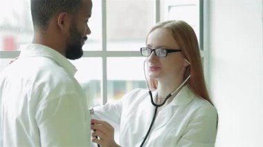 Parlak bir klinikte, gözlüklü, şefkatli bir kadın doktor genç bir adamın göğsünü nazikçe dinler ve bir anlık özenli bir sağlık hizmeti sergiler. Sıcak atmosfer, reçete yazmaya hazırlanırken profesyonel etkileşimlerini vurguluyor.