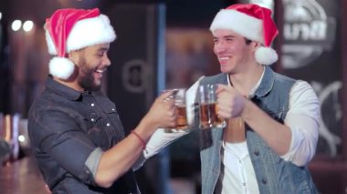 İki neşeli Noel Baba şapkalı çocuk, canlı bir bar atmosferinde neşe ve dostluk saçan soğuk bira bardaklarıyla kadeh kaldırıyor. Kahkahalar havayı dolduruyor. Arkadaşlarla çevrili tatil ruhunu kutluyorlar. Ve futbol maçının heyecanı devam ediyor.