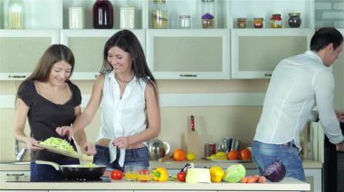 Parlak, modern bir mutfakta, üç genç arkadaş canlı salatalar hazırlıyor, renkli sebzeler doğruyor ve kahkahalarını paylaşıyorlar. Sahne neşe ve dostluk saçıyor, taze yeşillikler, sulu domatesler ve biraz da narenciye.