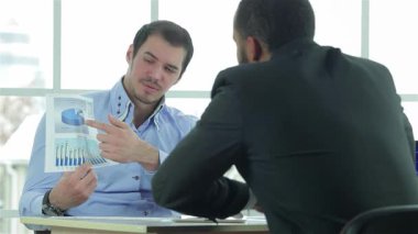 İki azimli iş adamı, resmi giyinmiş şık bir müzakere masasında renkli finansal tabloları analiz ederek dinamik bir tartışmaya giriştiler. Odaklanmış ifadeleri ve parlak ofis arkaplanı bir hırs ortamı yaratıyor.
