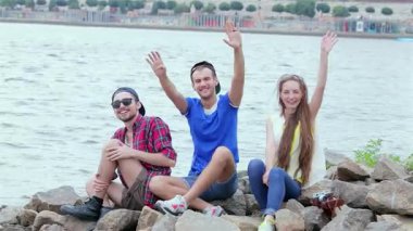 Üç neşeli arkadaş kayalık kıyılarda oturmuş, deniz kenarında güneşli bir günün tadını çıkarırken kameraya coşkuyla el sallıyorlar. Gülüşleri ve canlı gülümsemeleri arkadaşlığın ve kaygısız yaz maceralarının özünü yakalar.