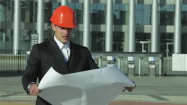 Gösterişli bir takım elbise ve parlak turuncu şapkalı odaklanmış bir mimar planları, hırsı ve profesyonelliği somutlaştırıyor. Modern inşaat alanı, şehir manzarasında yenilik ve takım çalışmasının bir karışımını sergileyerek potansiyelle uğulduyor.