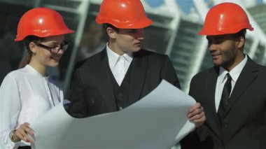 Üç kendine güvenen iş mimarı, bir kadın ve iki şık takım elbiseli ve parlak turuncu kasklı adam, bir inşaat sahasının planlarını dikkatle inceliyorlar. Onların odaklanmış ifadeleri ve takım çalışması yenilikçi yapı fikirlerinin heyecanını vurgular.