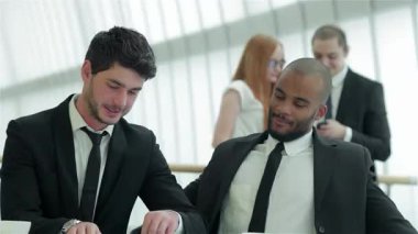 İki kendine güvenen işadamı şık bir ofis masasında belgeleri incelerken, arka planda iki iş arkadaşı da bir ışık anı paylaşıyor. Bu canlı sahne takım çalışmasının ve yeniliğin özünü yansıtıyor.