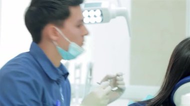 Mavi üniforma ve koruyucu maske takan odaklanmış genç bir dişçi, modern bir diş sandalyesinde hastayı muayene ederken ustalıkla diş aletlerini seçiyor. Sahne, profesyonel diş bakımının özünü yakalıyor ve...