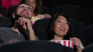 Neşeli bir çift, sıcak bir sinemada oturur, tamamen filmle meşguldürler, gülümsemeleri yüzlerini aydınlatır. İzleyiciler tarafından çevrelenmiş, patlamış mısır ve sodayı seviyorlar, paylaşılan heyecan ve eğlencelerden oluşan canlı bir atmosfer yaratıyorlar.