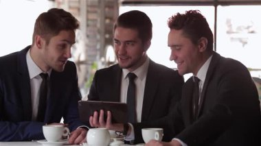 Üç iyi giyimli adam şık bir kafede iş yemeği sırasında canlı bir tartışmaya giriştiler, kahve ve tablet içerek fikir alışverişinde bulundular. Animasyon ifadeleri ve yoldaşlıkları başarılı bir ortaklığı, takım çalışmasını ve işbirliğini yansıtıyor.