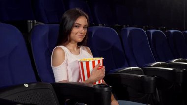 Çekici genç bir kadın kameraya gülümserken, lüks bir sinema koltuğunda çizgili patlamış mısır kovasını tutarken mutluluk saçıyor. Bu canlı sahne eğlence ve neşenin özünü yakalar, izleyicileri film büyüsünün heyecanını paylaşmaya davet eder.
