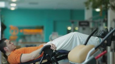 Orta Doğulu etnik kökene sahip odaklanmış genç bir adam, özel spor aletleri üzerinde sırt güçlendirme egzersizi yapıyor ve kararlılığını ve atletizmini sergiliyor. Canlı spor salonu, turkuaz duvarları ve modern aparatları var.