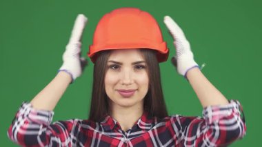 Yakın çekim bir stüdyo çekiminde kendine güvenen ve güzel bir kadın inşaat işçisini yakalıyor, canlı turuncu şapkasını düzeltirken pozitiflik saçıyor. Çekici gülüşü ve gösterişli ekose gömleği cesaret ve profesyonelliği temsil ediyor.