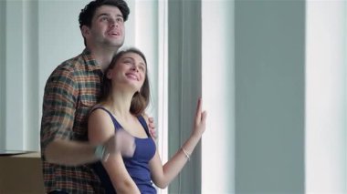 Neşeli bir çift, karton kutularla çevrili, parlak bir pencerenin önünde birlikte yeni bir hayat düşlüyorlar. Gülümsemeleri mutluluğu kucaklarken yeni evlerinde yeni bir başlangıç yapmanın heyecanını yakalıyor.