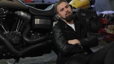 Şık siyah deri ceketli, sakallı genç bir adam güçlü motorsikletine yaslanarak düşünceli bir şekilde yerde oturuyor. Pırıl pırıl krom ve motorun karmaşık ayrıntıları biniciliğe olan tutkusunu yansıtıyor, bir macera ortamı yaratıyor.