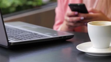Yakın çekim, sıcak bir kafe atmosferiyle çevrili cep telefonuna takılmış neşeli genç bir kadını yakalar. Sahne, bir fincan dumanı tüten kahve ve parlak bir dizüstü bilgisayarla büyütülmüş, gündelik bir haz ve bağlantı hissi uyandırıyor.