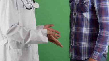 Kırpılmış bir çekim beyaz önlüklü bir erkek doktoru hastasıyla el sıkışırken yakalar. Bu da canlı yeşil renkli arka planda başarılı bir iyileşmeyi simgeler. Bu içten jest, sağlık hizmetlerinin doğasında olan güven ve desteği yansıtıyor.