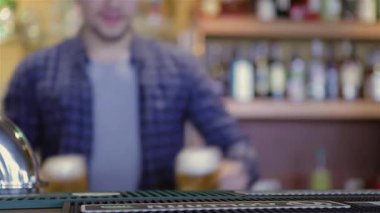 Yetenekli bir barmen, sıradan bir şekilde ekose gömlek giyer ve hareketli bir partide ustalıkla köpüklü iki bira servisi yapar. Canlı atmosfer renkli şişelerin arka planıyla güçlenir, konukları kutlama ve arkadaşlığın tadını çıkarmaya davet eder.