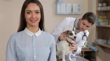 Neşeli bir Kafkasyalı kadın veteriner kliniğinde kendine güvenerek duruyor ve onay alabilmek için onay veriyor. Arka planda, yetenekli bir erkek veteriner küçük bir köpeği inceliyor, sıcak bir bakım ve profesyonellik atmosferi sergiliyor.