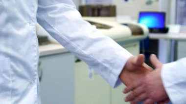Temiz beyaz önlüklü odaklanmış bir erkek bilim adamı sıkı bir tokalaşmadan sonra ellerini uzatarak laboratuvarda heyecanlı bir güne hazırlanıyor. Ellerinin birbirine masaj yapması, arka plandaki beklenti ve arkadaşlığı vurguluyor.