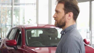 Neşeli, sakallı bir adam arabasının anahtarlarını çarpıcı kırmızı bir otomobilin önünde tutarken gururla ışıldıyor. Bu an, sahip olmanın heyecanını yakalıyor. Parlak, modern bir galeride yeni bir araç almanın sevincini gösteriyor.
