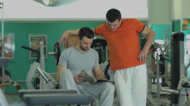 Canlı bir spor salonu ortamında, gri kıyafetli odaklanmış genç bir adam turuncu gömlekli spor eğitmeni bir spor dergisinden eğitim programını tartışmak için eğilirken oturuyor. Bu dinamik ikili adanmışlığı ve takım çalışmasını, teşhirciliği simgeler.