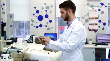 Gıcır gıcır beyaz laboratuvar önlüğü giyen kendini adamış genç beyaz bir bilim adamı özenle bilgisayardan parlak bir tablete veri giriyor. Bu sakallı stajyer, düşünceli bir ifadeyle gelişmiş laboratuvar ekipmanlarının arasında duruyor.