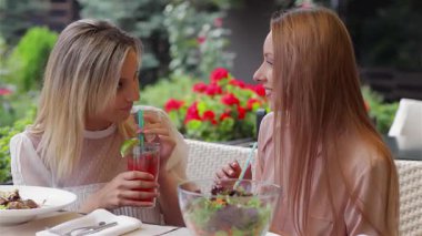 İki güzel kadın şık bir kafe terasında oturmuş renkli kokteyllerini yudumluyor ve canlı dedikodular yapıyorlar. Kahkahaları havayı dolduruyor, canlı çiçeklerle çevrili, neşeli bir arkadaşlık ve yaz eğlencesi ortamı yaratıyor.