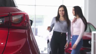 Parlak ve modern bir galeride, iki kadın parlak kırmızı bir araba hakkında animasyonlu bir tartışmaya girişiyor, ön planda ışıkları parıldıyor. Bu canlı sahne tüketiciliğin heyecanını ve yeni bir araç seçmenin neşesini yakalıyor.