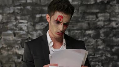 Gösterişli siyah takım elbiseli bir erkek vampir, öfkeyle bakan bir belge okuyor, onu koyu renkli tuğla bir duvara doğru parçalıyor. Dramatik makyajı, kana benzeyen çizgileri, ürkütücü Cadılar Bayramı atmosferini arttırır,