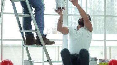 Parlak, havadar bir dairede merdivendeki gülümseyen bir adam oturmuş ortağına bir kutu boya uzatıyordu. Bu dinamik sahne, ev yenilemesindeki takım çalışmasının ve yaratıcılığın özünü yakalıyor ve bir alanı dönüştürmenin heyecanını gözler önüne seriyor.