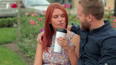 Neşeli bir çift parkta bir bankta yakın oturuyor, gülüşlerini paylaşıyorlar ve güllerin arasında kahve alıyorlar. Rahatlamış tavırları ve eğlenceli etkileşimleri açık hava ortamında sıcak bir dostluk ve bağlantı ortamı yaratıyor.