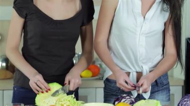 İki neşeli kız arkadaş neşeli bir şekilde canlı sebzeleri dilimlerken kameraya gülümsüyorlar ve sıcak ve davetkar bir ortam yaratıyorlar. Kahkahaları sıcacık mutfağı dolduruyor. Renkli ve sağlıklı bir yemek hazırlıyorlar.