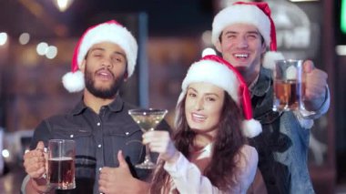 Neşeli bir üçlü, iki erkek ve bir kadın, neşeli Noel Baba şapkaları takıyorlar ve neşeli bir şekilde barlarında içkileriyle kadeh kaldırıyorlar. Sıcak atmosfer kahkahalarla ve dostluklarla dolu. Tatil ruhunu kutluyorlar.