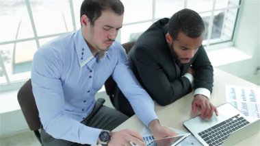 Doğal ışıkla yıkanan modern bir ofiste, iki odaklanmış işadamı tablet ve dizüstü bilgisayardaki verileri analiz ederken dinamik bir tartışmaya girişiyor. Resmi kıyafetleri ve ciddi ifadeleri, strateji oluştururken başarıya olan bağlılıklarını yansıtıyor.