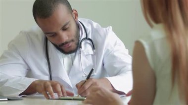 Şefkatli bir doktor parlak, modern bir klinikte reçete yazarken hastasını dikkatle dinler. Sıcak atmosfer, çok ırklı doktorla hasta arasındaki şefkatli ilişkiyi vurguluyor.