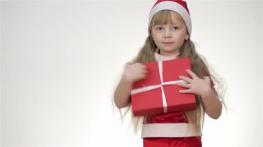 Şen şakrak giyinmiş ve Noel Baba şapkası takmış çekici bir kız bebek, yumuşak beyaz arka planda canlı kırmızı bir hediye kutusuyla poz veriyor. Neşeli ifadesi ve günümüzün etrafındaki nazik kucaklaşması tatil sezonunun neşesini ve mucizesini çağrıştırıyor.