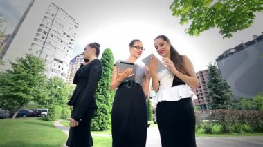 Kendine güvenen genç bir iş kadını modern bir ofis binasının önünde duruyor ve tabletini hazırlarken, iki meslektaşı yakınlarda sohbet ediyor. Canlı şehir arkaplanı profesyonellik ve işbirliği atmosferini artırıyor