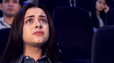 Uzun kahverengi saçlı, güzel beyaz bir kadın, koyu renkli bir tiyatroda oturuyor. Ekrandaki dramayı izlerken ifadesel suratı derin endişeleri yansıtıyor. Yakın çekim, onun yoğun duygularını yakalıyor, bulanık siluetlerle çevrili...