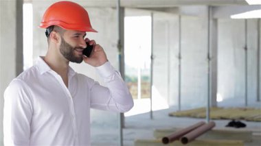 Canlı turuncu bir şapka takan genç bir Kafkas mühendisi, dinamik bir inşaat sahasının ortasında güvenli bir şekilde telefonla sohbet ediyor. Çekici bir gülümseme ve olumlu bir jest ile, başarıyı ve profesyonelliği somutlaştırıyor, hammaddeler ve