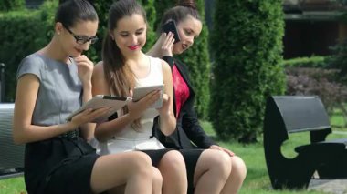 Üç kendine güvenen iş kadını parkta bir bankta oturmuş, cihazlarını kullanırken aktif olarak fikirleri tartışıyor ve paylaşıyorlar. Şık kıyafetleri ve canlı ifadeleri, yemyeşil şehir ortamında modern ve işbirlikçi bir ruhu yansıtıyor.