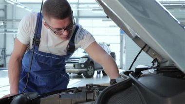 Odaklanmış, sarı saçlı beyaz bir tamirci açık kaputun üzerine eğilip motor parçalarını titizlikle inceliyor. Mavi tulum ve gözlük takarak profesyonelliği parlak ve modern bir garajda somutlaştırıyor.