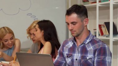 Yakışıklı sakallı bir erkek öğrenci dizüstü bilgisayarıyla meşgul olurken sevinçle ışıldıyor. Etrafı odaklanmış sınıf arkadaşlarıyla çevrili. Bu canlı sahne, modern bir öğrenme ortamında işbirliği ve coşkunun özünü yansıtıyor.