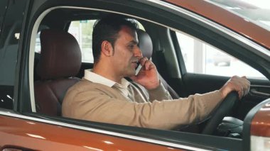 Koyu renk saçlı neşeli bir adam şık, modern bir arabada oturuyor ve canlı bir telefon görüşmesi yapıyor. Bej rengi süveteri, zengin kahverengi iç kısmıyla çok güzel zıtlaşır. Rahatlık ve tarz hissini yansıtır.