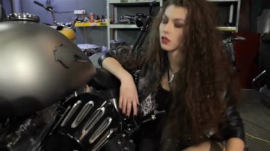 Göz kamaştırıcı bir bayan motorcu, özgüven ve cazibe saçarak zarif motorsiklet tankını nazikçe okşar. Kıvırcık saçlarıyla şık bir siyah deri ceketin üzerine dökülüyor. Canlı bir motosiklet atölyesinde özgürlük ve macera ruhunu simgeliyor.