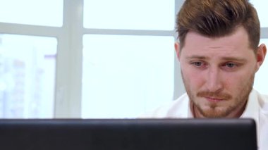 Yakışıklı, sakallı bir erkek yönetici parlak, çağdaş bir ofis alanı ile çevrili parlak dizüstü bilgisayarına dikkatlice yazı yazar. Beyaz gömleği, iç kısmın sıcak tonlarıyla çelişiyor. Hırsla dolu profesyonel bir atmosferi yansıtıyor.