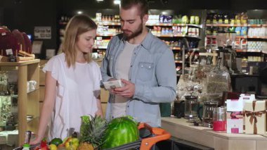 Çekici genç bir çift renkli bir hipermarketi inceliyor, dikkatle çay paketini inceliyor. Çekici beyaz adam etiketi okurken kadın gülümsüyor ve ortak bir keşif ve neşe ortamı yaratıyor.