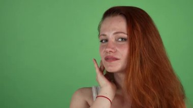 Dalgalı kızıl saçlı çarpıcı genç bir kadın neşeli bir şekilde yeşil renkli bir arka plana karşı poz verirken neşe saçıyor. Yüzündeki nazik dokunuşu ve neşeli gülümsemesi, sıcaklığı ve güveni çağrıştırıyor, izleyicileri kucaklamaya çağırıyor.